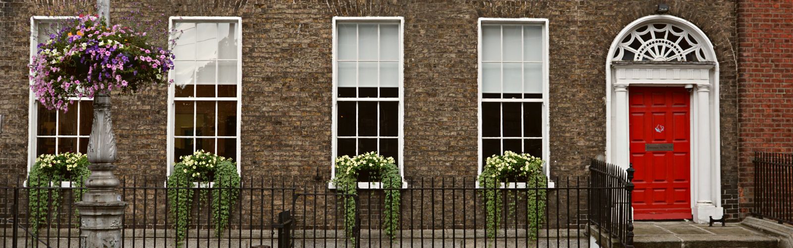 The exterior of a brown stone house, with four windows each carrying a potted green plant. To the right of the windows, there is a red door trimmed with white. 